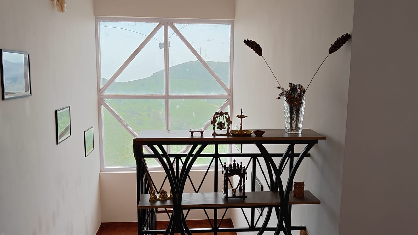 A console table of brass pieces and dried flowers below the latticed landing window, hills filling the frame behind