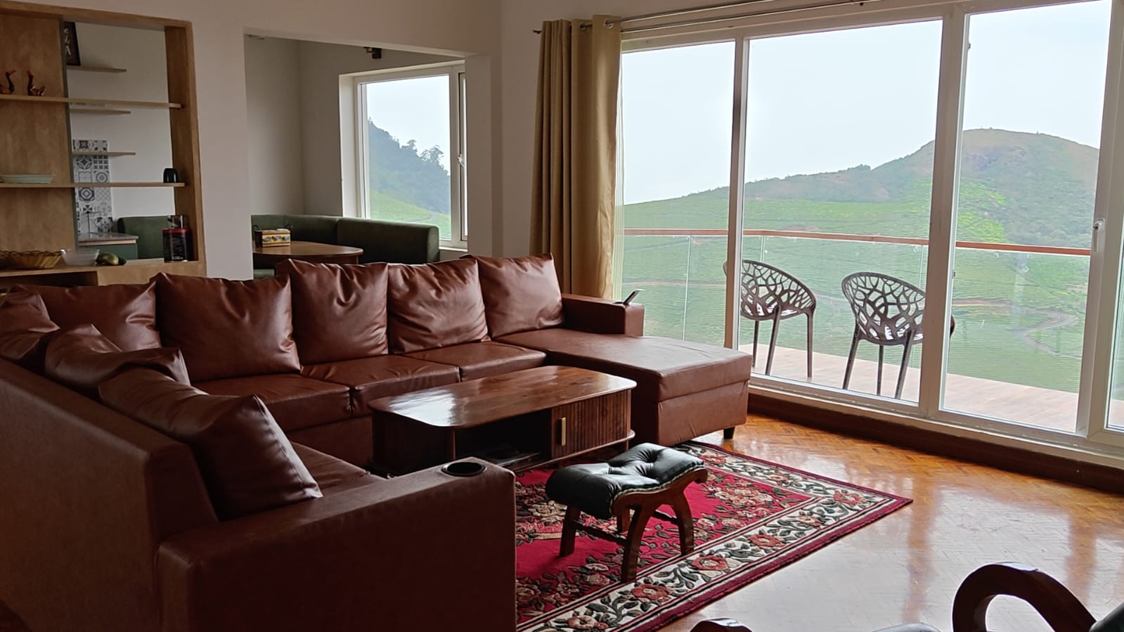 The living room looking out through full-height glass to the balcony chairs, with the tea garden hills beyond